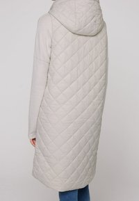 Vadderad ljusbeige väst med huva. Mjuka tröjärm, sömlös design och en smickrande längd. Har ett diamantmönster på tyget.