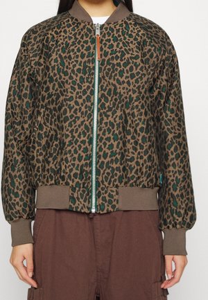 Leopard print bomber jacket with green accents, ribbed collar and cuffs, silver zipper, worn over white top and brown pants.