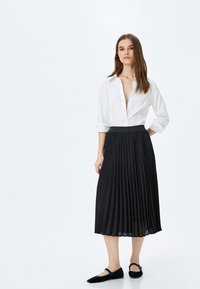 Falda midi negra con pliegues y una cinturilla suave, combinada con una camisa blanca de cuello. La modelo lleva zapatos planos negros. Diseño simple y moderno.