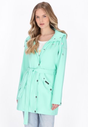 Młoda kobieta z długimi blond włosami, ubrana w miętowozielony płaszcz przeciwdeszczowy z paskiem, na białej bluzce i jasnoniebieskich dżinsach, na jednolitym tle.