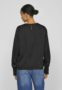 Blusa nera a maniche lunghe con dettaglio a chiave sulla schiena. Tessuto morbido, vestibilità comoda, dettagli testurizzati ai polsini delle maniche. Indossata con jeans blu.