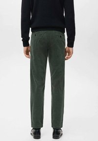 Mężczyzna ubrany w ciemnozielone spodnie sztruksowe, czarny sweter i czarne eleganckie buty, widziany od tyłu na jasnym, jednolitym tle.