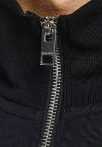 Musta vetoketjullinen takki, jossa on ribattu pinta, korkea kaulus ja hopeinen vetoketju suoralla vetotapilla, jossa lukee "JACK & JONES".