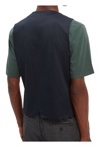 Uomo che indossa un gilet senza maniche blu navy sopra una camicia verde a maniche corte e pantaloni scuri a quadretti, visto da dietro su uno sfondo bianco.