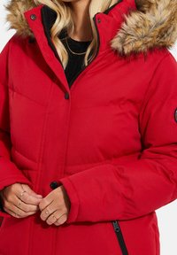 Doudoune rouge avec une capuche à fausse fourrure, dotée d'une fermeture éclair, de boutons sur le devant et d'une texture matelassée. Des mains ajustent la doudoune.