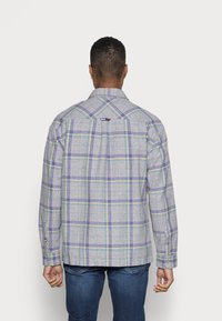Uomo che indossa una camicia a maniche lunghe grigia a quadri con linee viola, gialle e blu, abbinata a jeans blu scuro, visto da dietro.