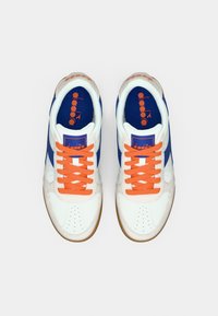 Λευκά αθλητικά παπούτσια με μπλε λεπτομέρειες, πορτοκαλί κορδόνια και σόλα από καουτσούκ. Διαθέτουν οπές στο μπροστινό μέρος και λογότυπο στη γλώσσα.