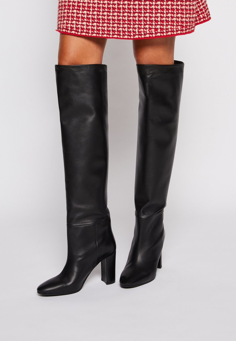 Nohy v černých kožen�ých vysokých botách na blokovém podpatku, spárované se sukní s červeným a bílým vzorem, na jednoduchém pozadí.