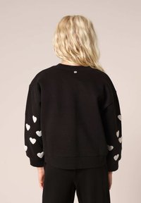 Zwarte sweater met zilveren hartborduursel op de mouwen, geribbelde manchetten en zoom. Eenvoudige ronde halslijn en een relaxed fit.