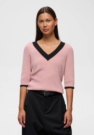 Femme aux longs cheveux foncés portant un pull rose à col en V avec bordure noire et un pantalon noir avec ceinture, debout devant un fond uni.