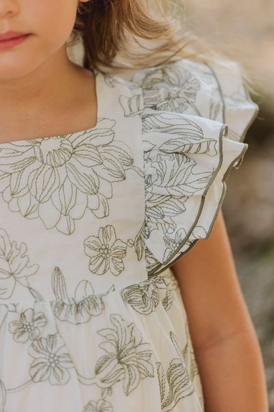 Vit klänning med grön blommig broderi och volangärmar. Har en fyrkantig halsringning och en rynkad kjol. Texturerad tyg med subtil lyster.