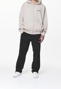 Sudadera con capucha beige claro con cremallera, pantalones cargo negros y zapatillas blancas. La sudadera tiene un bolsillo frontal y un logo bordado. Los pantalones cuentan con bolsillos laterales.