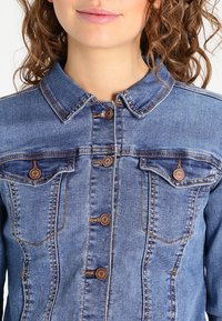 Chaqueta de denim con cuello, botones marrones y dos bolsillos en el pecho. La tela es azul con un sutil efecto de desgaste y costuras visibles.