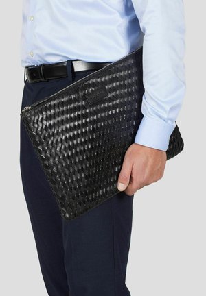 Mann i blå skjorte og svarte bukser som holder en svart flettet lærveske med glidelås ved siden av seg.