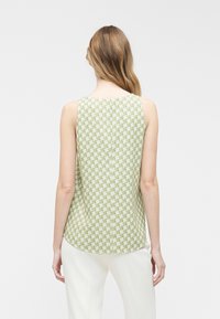 Femme aux longs cheveux portant un haut sans manches vert clair et blanc à motifs et un pantalon blanc, vue de dos sur un fond uni.