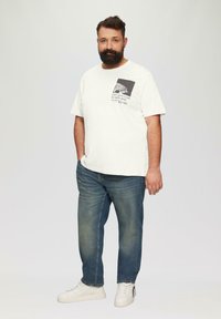 Witte graphic t-shirt met een borstprint, gecombineerd met middelblauwe jeans en witte sneakers. Het shirt heeft een comfortabele pasvorm en korte mouwen.