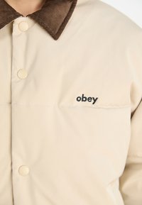 Bež jakna s podstavom i okovratnikom od kordura smeđe boje. Ima prednje snape i crni "obey" logo na grudima. Glatka tekstura.