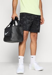 Μαύρη τσάντα duffel με λευκό λογότυπο Nike, φορεμένη με μαύρα παντελόνια καμουφλάζ, λευκές κάλτσες και λευκά αθλητικά παπούτσια. Ανοιχτό πράσινο αθλητικό πουκάμισο.