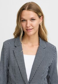 Fekete-fehér houndstooth mintás blézer, illeszkedő fazonnal, kihajtott gallérral és textúrált anyaggal, fehér egyszínű felsőre viselve.