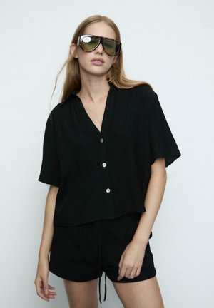 Junge Frau trägt übergroße getönte Sonnenbrillen, ein lockeres schwarzes kurzärmeliges Hemd mit Knöpfen und passende schwarze Shorts vor einem schlichten Hintergrund.