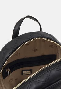Intérieur d'un sac à dos en simili cuir noir matelassé avec une poche zippée et une doublure beige arborant un motif de logo répétitif. Détail des fermetures éclair en or.