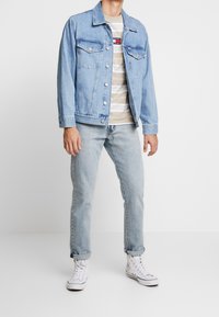 Uomo che indossa una giacca di jeans azzurro chiaro sopra una camicia beige a righe, jeans azzurri con orli e sneaker bianche alte, in piedi su un pavimento bianco.