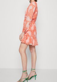 Korallefarbenes Kleid mit Paisleymuster, langen Ärmeln und Rüschensaum. Kombiniert mit grünen Riemchenschuhen. Glatter Stoff, leichtes Material.