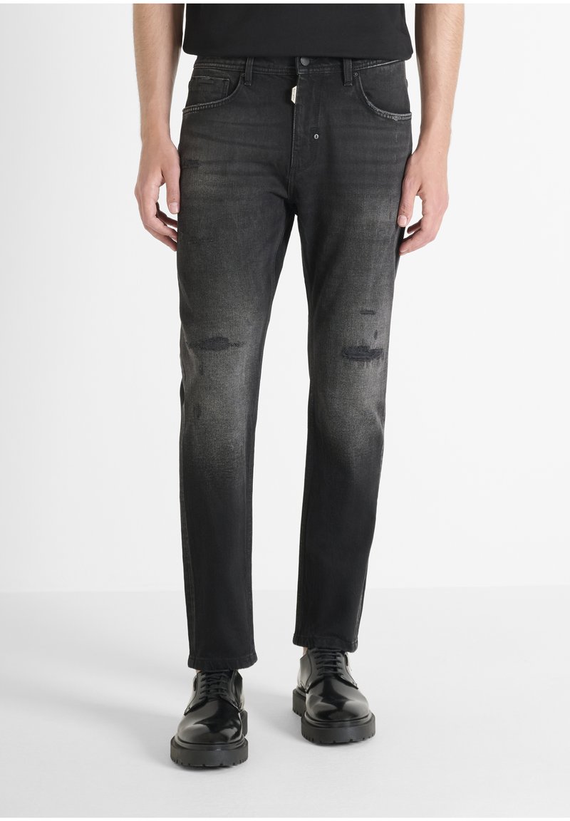 Vaqueros de denim negro con áreas desgastadas y detalles deshilachados. Diseño de pierna recta, estilo de cinco bolsillos, combinados con zapatos negros pulidos.