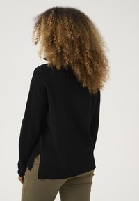 Schwarzer strukturierter Pullover mit hohem Kragen, seitlichen Schlitzen und entspannter Passform, kombiniert mit olivgrünen Hosen. Fokus auf Einfachheit und Komfort.