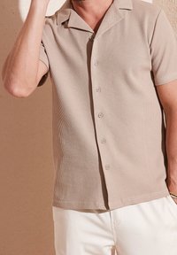 Beige kortärmad skjorta med en strukturerad mönstring, spridd krage och knappar fram, i kombination med vita byxor.