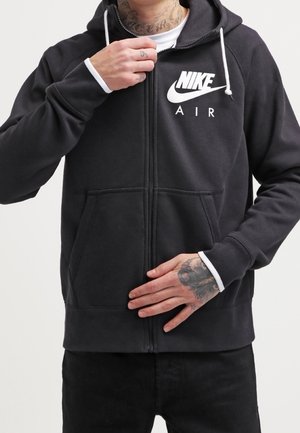 Person wearing black Nike Air zip-up hoodie, hand adjusting zipper, visible tattoo on neck and hand, white drawstrings on hood.