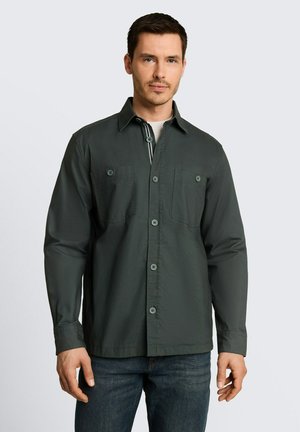 Man wearing a dark green button-up shirt with chest pockets and blue jeans standing against a light gray background.