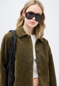 Νεαρή γυναίκα που φοράει υπερμεγέθεις μαύρα γυαλιά ηλίου, ελαινοπράσινο σακάκι με κουμπιά, λευκή μπλούζα, ασημένια σκουλαρίκια κρίκους και κρατά μια μαύρη, υφασμάτινη τσάντα ώμου.