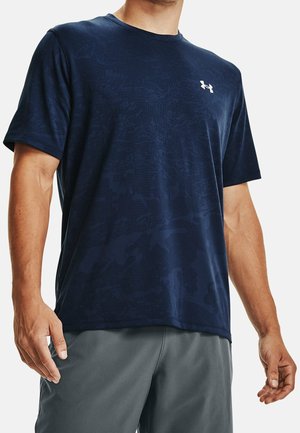Man draagt een donkerblauw Under Armour T-shirt met korte mouwen en een subtiel patroon, en grijze korte broek, staat met ontspannen armen langs het lichaam.