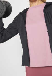 Tenue de sport comprenant un sweat à capuche zippé noir, un t-shirt rose et un legging rose ; comprend un haltère dans la main avec une prise texturée.