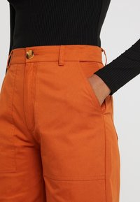 Oranje cargobroek van katoen, voorzien van zijzakken, een rechte pasvorm en een gouden knoop bij de tailleband, gecombineerd met een zwart geribd topje.