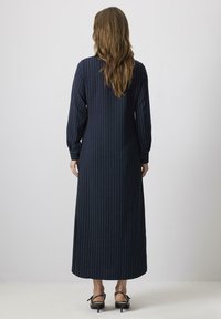 Rochie lungă de culoare bleumarin, cu dungi verticale albe, mâneci lungi și o siluetă dreaptă. Purcată cu sandale negre cu bretele.