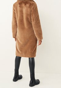 Manteau en fausse fourrure brun, ébouriffé, avec un col, des manches longues et une silhouette droite. Associé à des legging noirs et brillants ainsi qu'à des bottines.