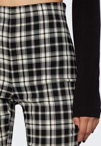 Gros plan d'une personne portant un pantalon à carreaux noir et blanc et un haut noir à manches longues, la main reposant le long du corps.