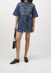 Denim dress with short sleeves, belted waist, and button-down front. Dark blue color, two chest pockets, and frayed hem detail. Black mules and bag.