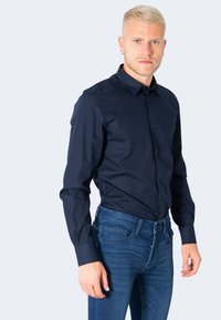 Camicia azzurra scuro a maniche lunghe, in taglio slim, realizzata in tessuto liscio. Presenta un colletto classico e una chiusura con bottoni; indossata con jeans in denim.