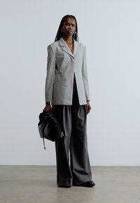 Γκρι σακάκι με κομψές πέτο και ένα κουμπί, συνδυασμένο με άνετο γκρι παντελόνι. Κρατάει μια μαύρη δερμάτινη τσάντα με κορδόνι.