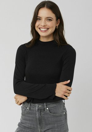 Haut noir à manches longues avec un col montant, fabriqué dans un matériau texturé. Associé à un jean gris et à de petites boucles d'oreilles dorées minimalistes.