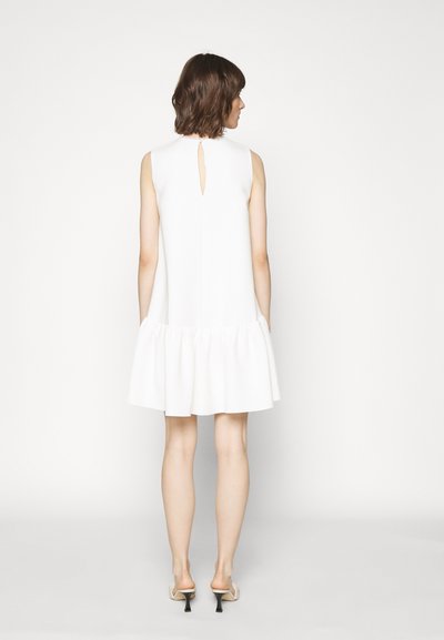 Bjela haljina bez rukava s lepršavim rubom, detaljem ključanice na leđima i glatkom teksturom, doseže sredinu bedara. Nosiva s cipelama svijetle boje otvorenih leđa.