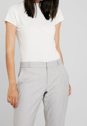 Kobieta ubrana w białą bluzkę z krótkim rękawem wpuszczoną w jasnoszare, dopasowane spodnie, na tle jednolitego jasnoszarego tła.