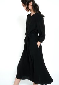 Žena s dugom tamnom kosom u crnoj midi haljini dugih rukava i svezanom strukom, pozira bočno s jednom rukom u džepu.