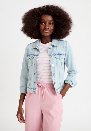 Frau mit voluminösem lockigem Haar, die eine helle Jeansjacke über einem rosa gestreiften Hemd und rosa Hosen mit Kordelzug trägt, steht mit einer Hand in der Tasche.