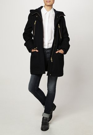 Manteau noir avec capuche, doté de fermetures éclair dorées, deux poches latérales, manches longues et coupe décontractée, porté sur une chemise blanche à col et un jean.