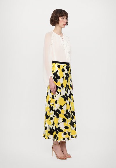 White blouse with ruffle detailing paired with a long yellow skirt featuring a black and white floral pattern. Tan high-heeled sandals.