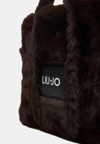 Borsa a tracolla in pelliccia sintetica marrone con forma strutturata, caratterizzata da una toppa con logo nero. Texture morbida e doppi manici per il trasporto.